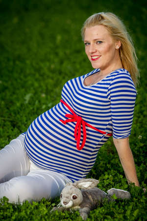 A cute pregnant girl with a striped top sitting in a green grass holding a fluffy bunny toy.,の写真素材