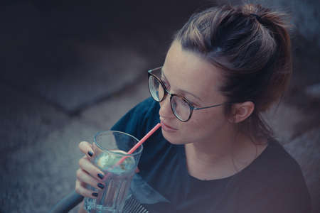 Young sexy caucasian hipster woman with glasses holding a glass with gin tonic drink and sipping it through a pink straw. Home garden atmospehere in evening-の写真素材