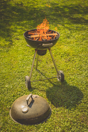 Round mobile grill or barbecue parked on a green surface or lawn. Visible flame and fire on a wooden and coal fuel. The grille is empty of meat and the lid is resting on the grass.の写真素材