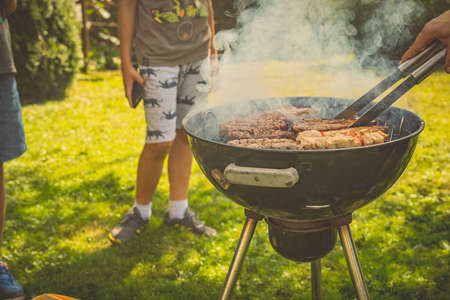 Kids waiting to receive meat from a grill while waiting behind it. Round grill in smoke with cevapcici on it on a garden lawn with a hand turning meat.の写真素材