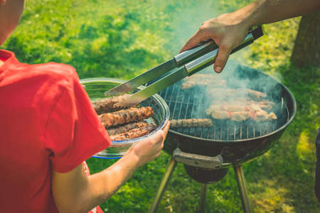 Unknown kid in a red shirt is recieving meat from a grill while holding a glass plate or bowl with ready meat. Round grill in smoke with cevapcici on it on a garden lawnの写真素材