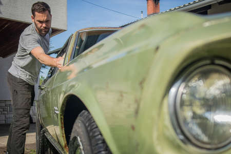 Young man with a hip beard cleaning a window of an old green vintage car on a home lawn. Cleaning muscle car windows at home with a cloth.の写真素材