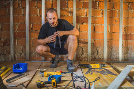 Man kneeling on a construction of a roof, holding tin drywall holders and thinking which tool will he use for the job. man caught between different work appliances.の写真素材