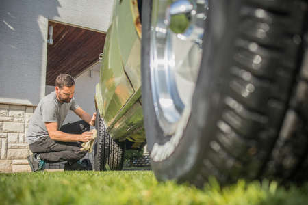 Young man with a hip beard cleaning a wheel of an old green vintage car on a home lawn. Cleaning muscle car tires at home with a cloth.の写真素材