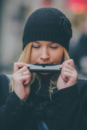 Frontal portrait of young attractive blonde woman playing harmonica on the street. Woman wearing a black hat, jacket and playing a harmonica with her mouth with eyes closed.の写真素材
