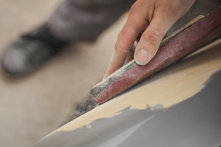 A hand of a man is seen sanding an old vintage car using a block of wood and brown sand grit paper. Manual dry sanding of a car in restoration, a process where putty is removed from bodywork.の写真素材