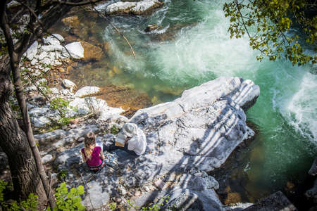 A couple sitting outside on a rock on a riverbank, enjoying the shade and reading a book next to a wild green river with rapids.の写真素材