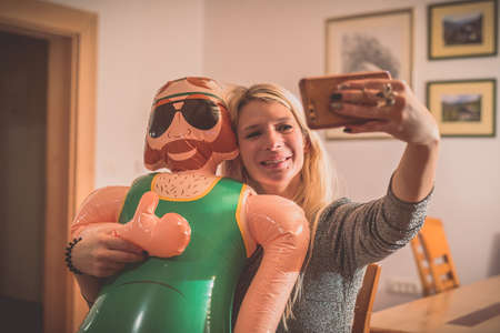 A smiling young blonde woman making a selfie wih a smartphone with an inflatable male doll. Party environment inside with a funny game and inflatable doll.の写真素材