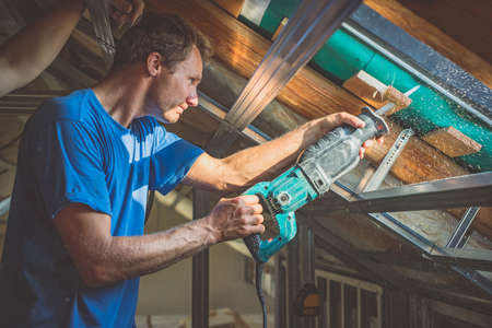 A person is cutting a board of wood next to a window with a reciprocating saw. Sawdust is flying around. Detail of a man cutting wooden board with strong back light to emphasize saw dust.の写真素材