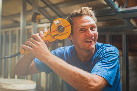 A young caucasian man is trying to cut his head open with an angle grinder. Frustration during home renovation or improvement, but still keeping a big smile on his face.の写真素材