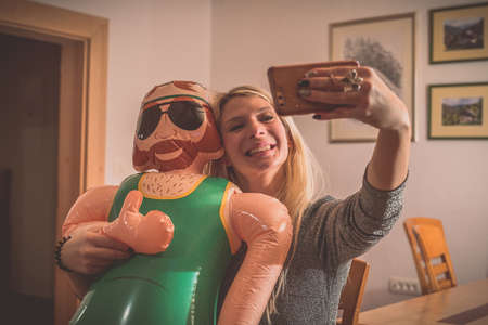 A smiling young blonde woman making a selfie wih a smartphone with an inflatable male doll. Party environment inside with a funny game and inflatable doll.の写真素材
