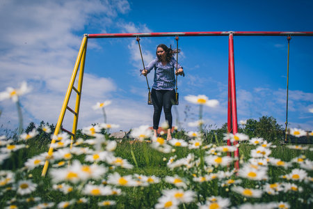 Young brunette woman is swinging on a swing, with a field of daisies (anthemidae) in the foreground on a beautiful spring sunny day. Concept of natural playfulness outdoor.の写真素材