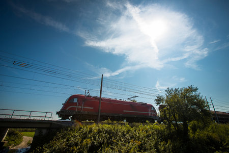 Modern european locomotive in red color is hauling a freight train over an embankment on a sunny day.の写真素材