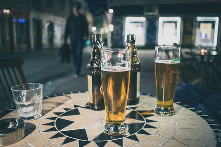 Two lager beers in half litre glasses on a bar table outdoor during evening hours, with brown bottles behind. A person is walking towards the table on a street at night.の写真素材