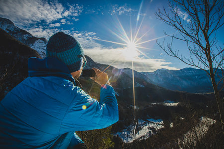 Strong lens flare with a hiker taking a photograph of a valley on alpine mountain. Sun is making a strong flare above the alpine valley.の写真素材