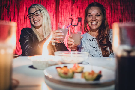 Two young women, a blonde and a redhead are cheering with glasses full of gin tonic drink and straws on a garden pary with candle on early eveningの写真素材