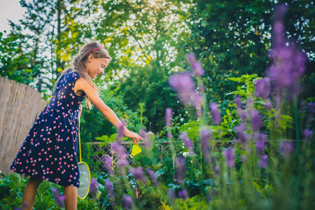 Young caucasian girl or kid is playing badminton game in a lush green garden. Kid holding shuttlecock and is preparing to serveの写真素材