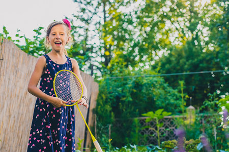 Young caucasian girl or kid is playing badminton game in a lush green garden. Kid enjoying in a garden while holding a racketの写真素材