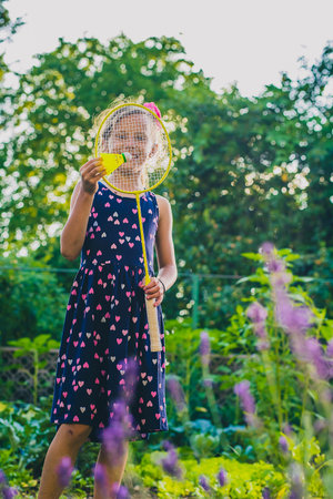 Young caucasian girl or kid is playing badminton game in a lush green garden. Kid holding a badminton racket in front of faceの写真素材