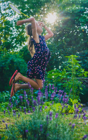 Young caucasian girl or kid is playing badminton game in a lush green garden. Kid jumping high up into sun while playing badmintonの写真素材