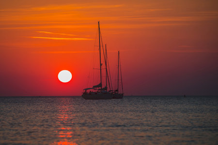 Bloody red sun setting down behind the silohouettes of a two sailboats or yachts moored on an open sea. Adventourous sunset.の写真素材