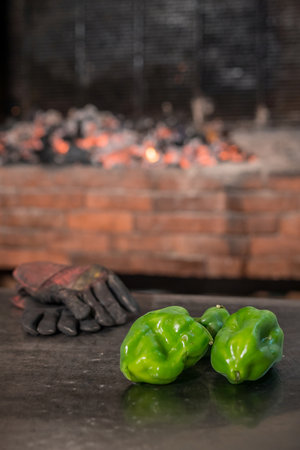 Two vegetable green paprikas and a pair of gloves as somewhat essentials of a good grill. Heating up charcoal grill in the background.の写真素材