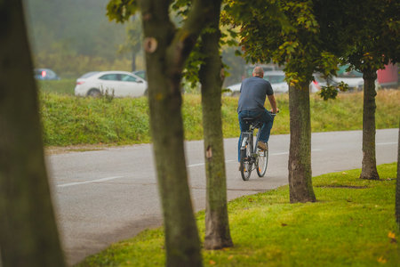 Bald man in jeans on a blue bicycle rushing towards work in a misty foggy morning commute on a small road with a row of trees. Cars in the background.の写真素材