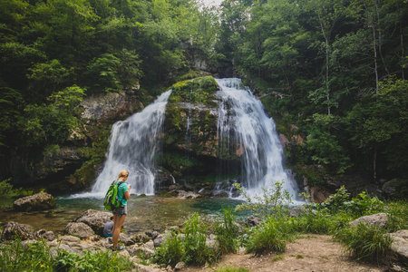 Blonde woman looking towards Virje waterfall in Slovenia, close to bovec. Dreamy and enchanting water falling down the cliff above the small alpine lake.の写真素材
