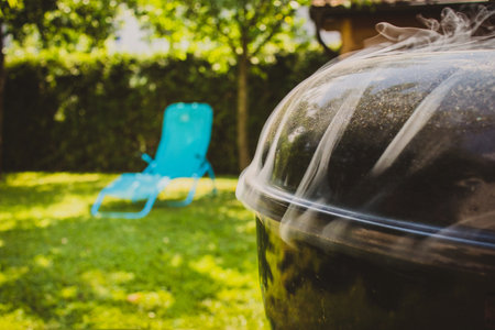Smoker BBQ grill with closed lid and smoke slowly coming up at the side. Seat or deckchair in the background. Concept of relaxation and barbecue.の写真素材