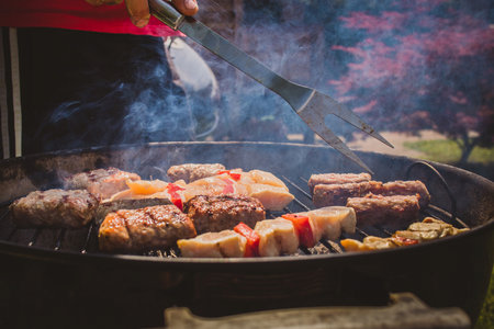 An assortment of meat like cevapcici, sausages and chicken kebabs on a charcoal grill. Green grass and maple in the background.の写真素材