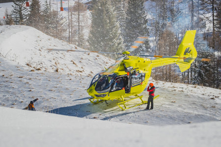 A yellow rescue helicopter is parked in the middle of the ski slope on a patch of snow. Concept of helicopter rescue on snow or ski slope. Visible pilots and other people around the helicopter.の写真素材