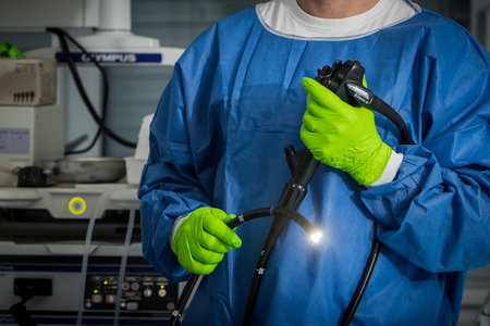 Doctor in blue protective clothing with green gloves is holding an endoscope in hand. The light on the endoscope is on, background with medicine utensils and instruments.の写真素材