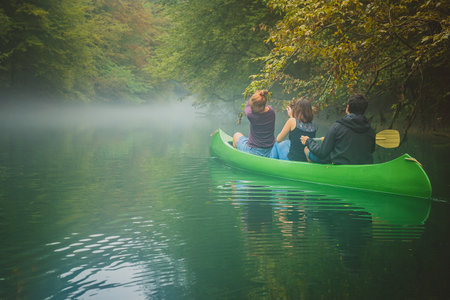 Group of three people, two young caucasian women and a man exploring, paddling with green canoe in misty and cold water with fog and green background.の写真素材