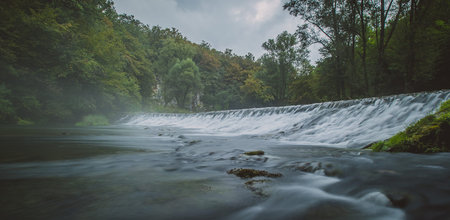 Panorama of river source or spring of Krupa in Bela Krajina (White Carniola) in Slovenia on a misty cloudy day. Foggy green river with rock formation in the background and fast water flowing over a long mystical dam and milky water.の写真素材