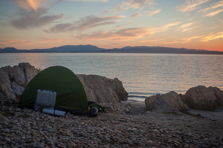 Wildcamping on the beach on an early morning. Tent pitched on stones next to the sea, with the sun just rising up from behind the hills.の写真素材