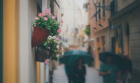 Romantic rainy busy street or alley with pots with flowers hanging on the wall . People with umbrellas walking past on the street.の写真素材