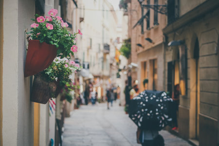 Romantic rainy busy street or alley with pots with flowers hanging on the wall . People with umbrellas walking past on the street.の写真素材
