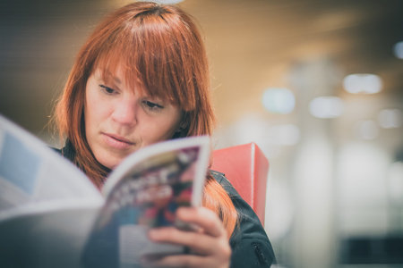 A young red haired woman is sitting in the airport and holding a travel book or tourist guide in her...