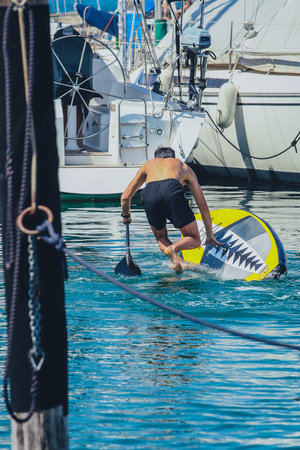Back view of a man falling from a SUP board into the sea in a marina between parked or moored boats. Goofy acting on a sup board sequenceの写真素材