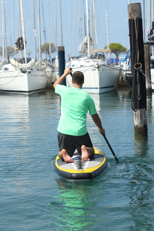 Young man kneeling on a SUP board and paddling around in a marina with parked boats around on a sunny day.の写真素材