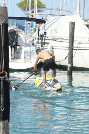 Back view of a man falling from a SUP board into the sea in a marina between parked or moored boats. Goofy acting on a sup board sequenceの写真素材