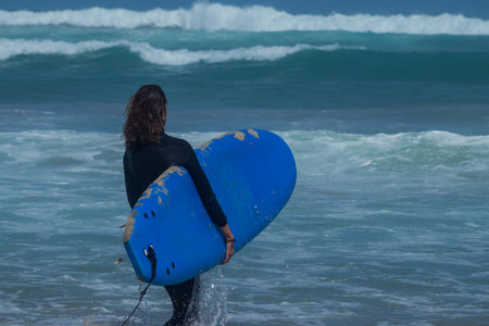 Back view of a surfer woman with long brown hair, holding blue surfboard and entering water with some waves in Dakar, Senegal.の写真素材