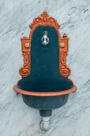 Front view of a vintage ornamental water basin and faucet mounted on a marble wall. Golden ornaments...