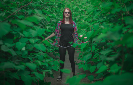 Young woman standing in the middle of the green bushes or shrubbery with sunglasses.