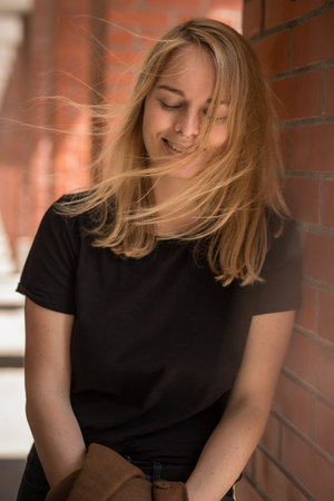 Portrait of young woman leaning against a brick wall, wearing black shirt and has long blonde hair that is blowing in the wind, the overall mood of the image is peaceful and serene.の写真素材