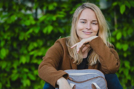 Young woman sitting on a grassy lawn with a backpack, brown jacket, black pants, background is garden or park with bushes. the overall mood of the image is peaceful and relaxed.の写真素材