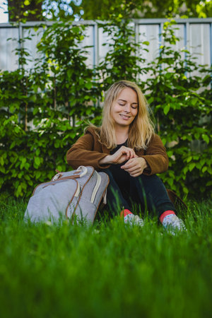 Young woman sitting on a grassy lawn with a backpack, brown jacket, black pants, background is garden or park with bushes. the overall mood of the image is peaceful and relaxed.の写真素材