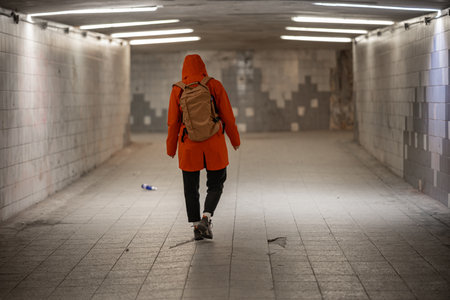A person in an orange coat with a backpack walks through a tiled underpass in Tallinn. The tunnel is lit by overhead lights, with a stark, empty atmosphere, reflecting urban solitude.の写真素材