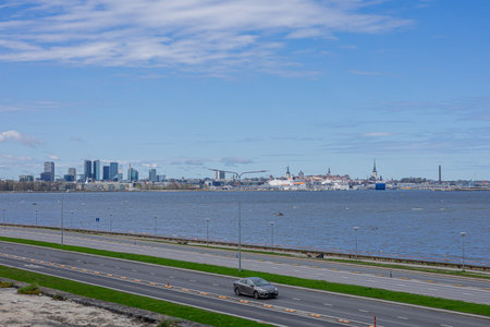 scenic view of Tallinns coastline along the Baltic Sea, with a highway running parallel to the water. The city skyline, featuring modern skyscrapers and historic spires, rises against a partly cloudy blue sky. Cars drive along the road, with the sea stretching toward the horizon.の写真素材