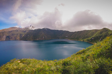The image shows Cuicocha Lake, a stunning crater lake in Ecuador, nestled within a volcanic caldera. Deep blue waters reflect the sky, surrounded by steep, green hills and distant mountains under a cloudy skyの写真素材